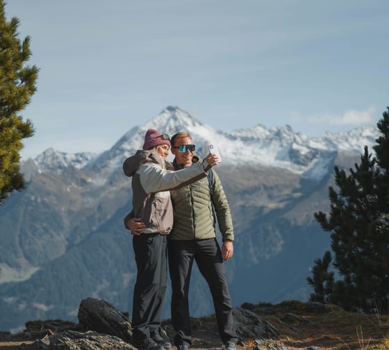 Ein Paar beim Wandern in den Bergen, umgeben von Tannenbäumen und schneebedeckten Gipfeln, macht ein Selfie.