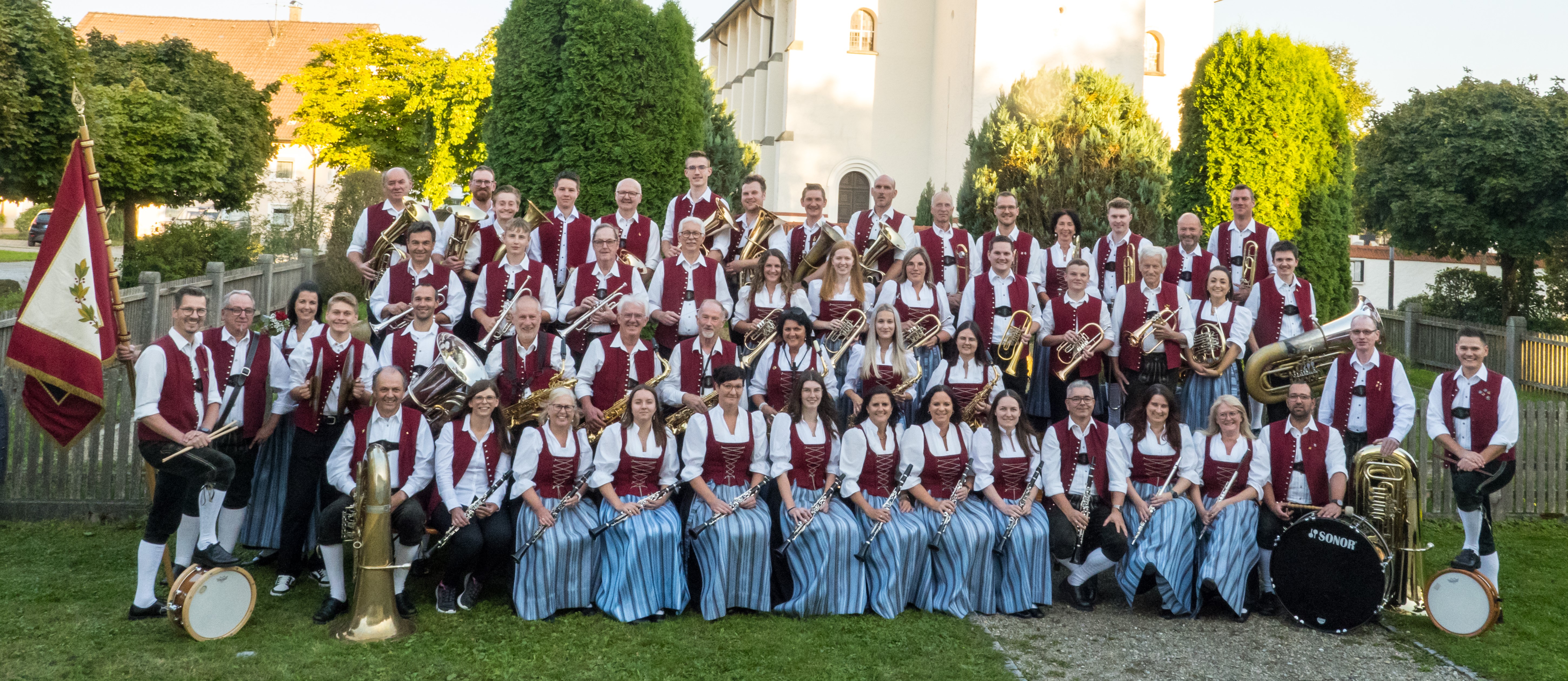 Das Bild zeigt eine große Blaskapelle in traditioneller Tracht, die vor einer Kirche für ein Gruppenfoto posiert, mit verschiedenen Blasmusikinstrumenten sowie Schlagzeug.