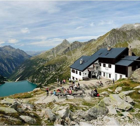 A mountain lodge with a black roof, surrounded by mountains and a lake, with visitors outside.