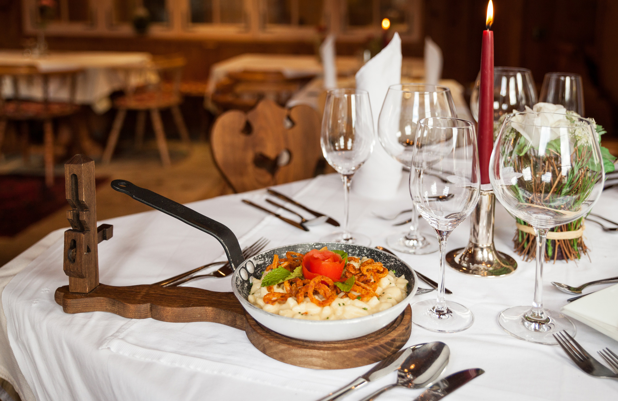 Käsespätzle mit goldbraunen Röstzwiebeln in einer Pfanne auf einem Holzbrett serviert. Der Tisch ist weiß gedeckt mit Weingläsern und einer brennenden roten Kerze im Hintergrund des gemütlichen Gasthofs Brücke.