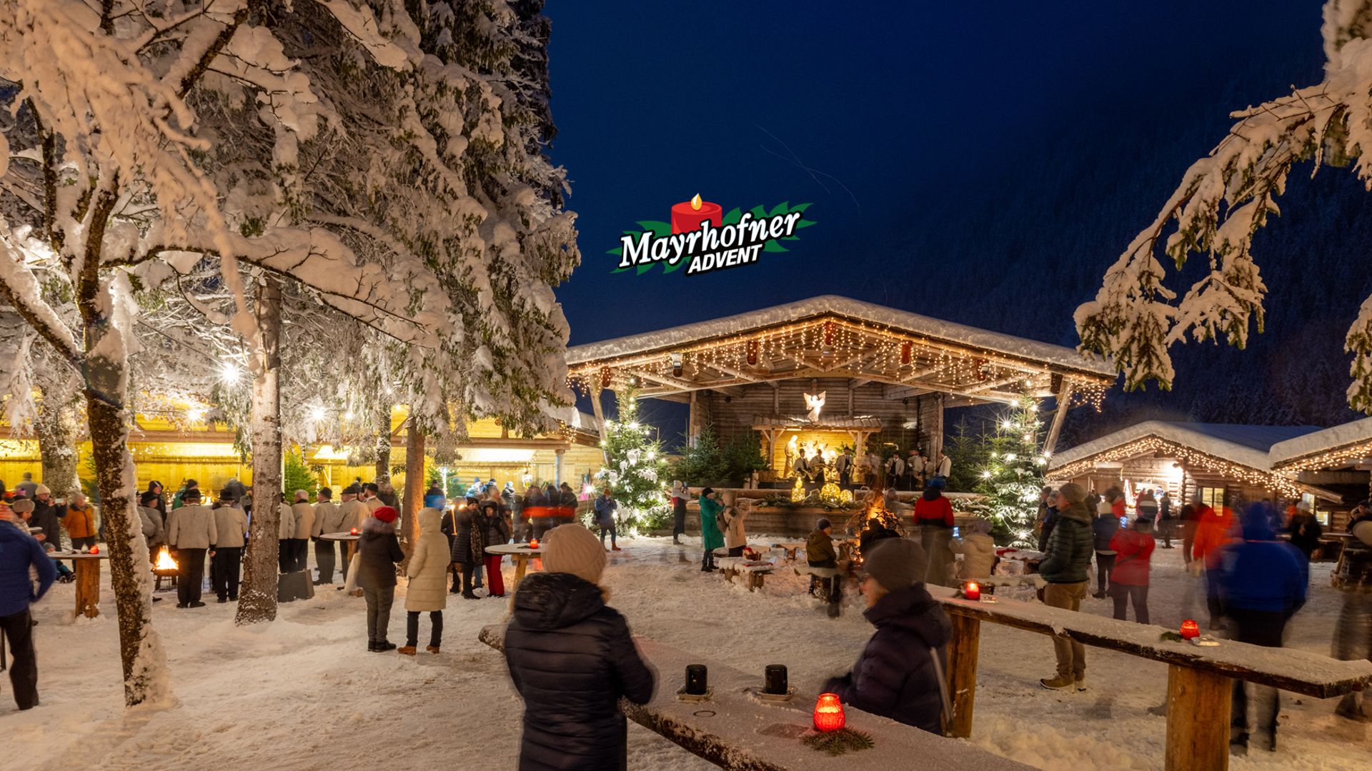 Winterliche Szene am Waldfestplatz in Mayrhofen: Menschen genießen den Mayrhofner Advent zwischen schneebedeckten Bäumen, Lichterketten und festlich beleuchteten Hütten. Im Hintergrund erstrahlt das große „Mayrhofner Advent“-Schild.