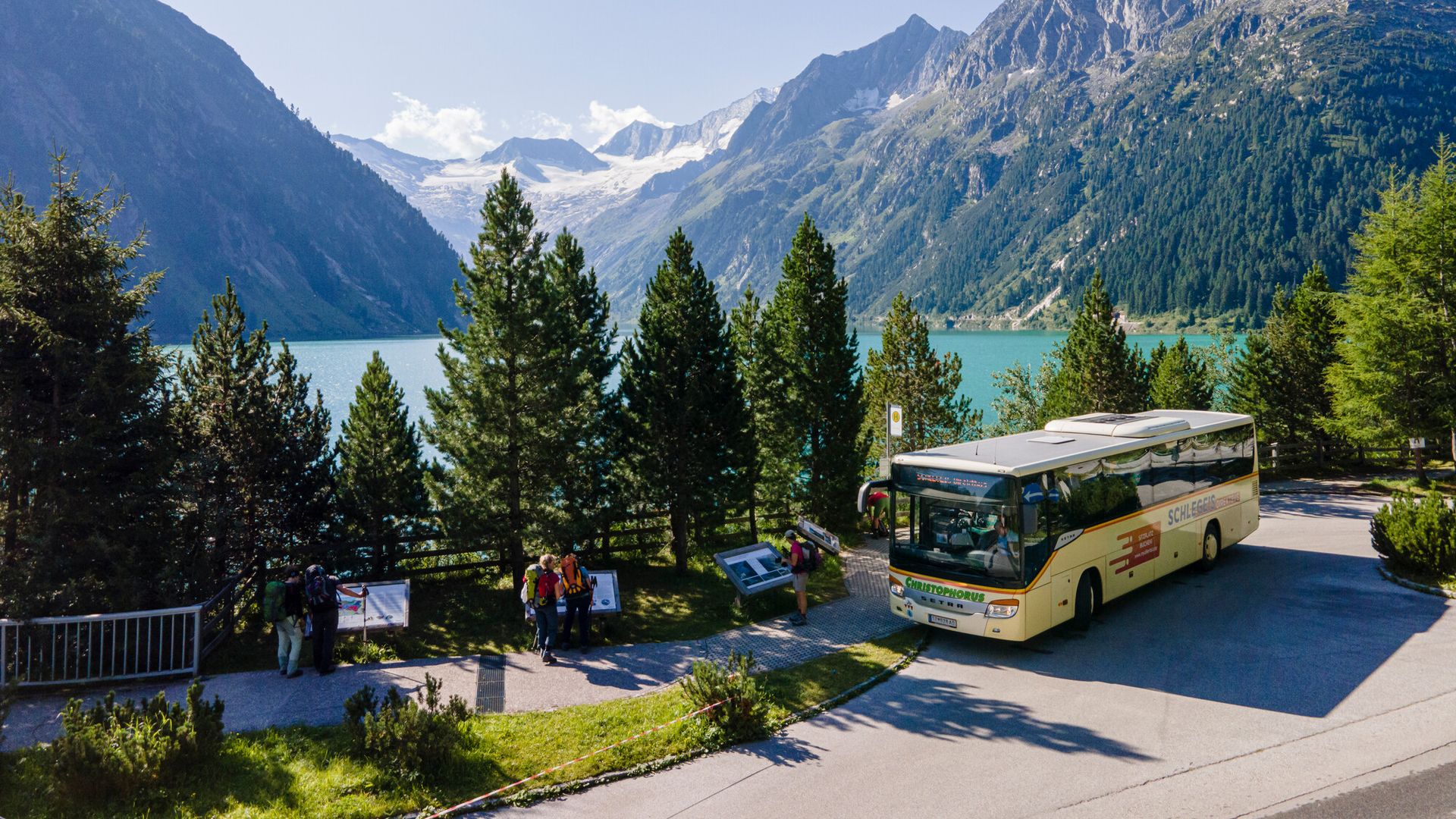 Ein gelber Linienbus steht an der Haltestelle am Schlegeisspeicher im Zillertal. Umgeben von grünen Bäumen, mit Blick auf den türkisfarbenen See und hohe Berge. Im Hintergrund schneebedeckte Gipfel unter blauem Himmel.