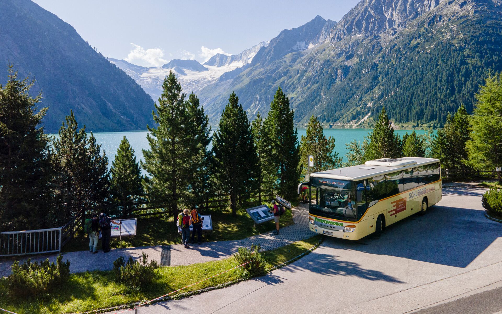 Ein gelber Linienbus steht an der Haltestelle am Schlegeisspeicher im Zillertal. Umgeben von grünen Bäumen, mit Blick auf den türkisfarbenen See und hohe Berge. Im Hintergrund schneebedeckte Gipfel unter blauem Himmel.