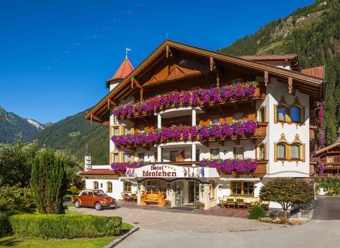 The image shows the exterior view of the Hotel Edenlehen with many flowers on the balcony. In front of the hotel, there is a car, a table with 4 chairs, and a few bushes.