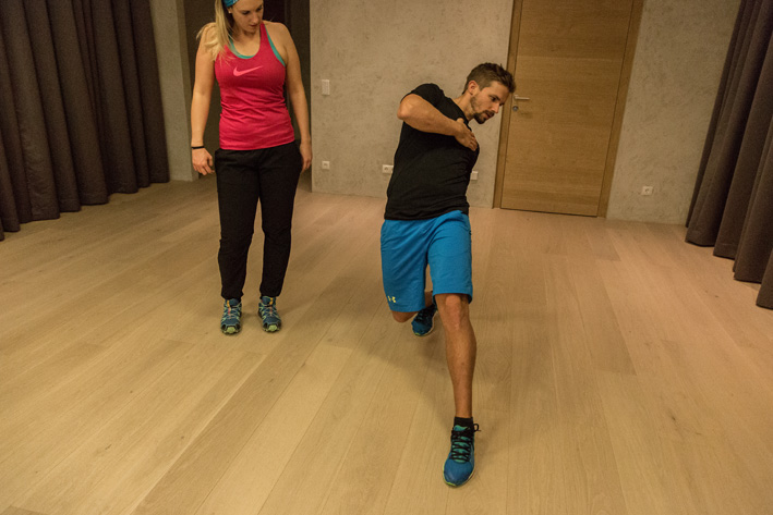 Mann führt diagonale Dehnübung im Trainingsraum in Mayrhofen aus, beobachtet von Trainingspartnerin. Christoph Ebenbichler zeigt Bewegung zur Verbesserung von Balance, Mobilität und Koordination für Skifitness.