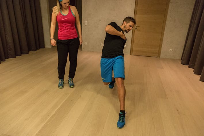 Man performing diagonal stretch exercise in training room in Mayrhofen, observed by training partner. Christoph Ebenbichler demonstrates movement to improve balance, mobility and coordination for ski fitness.