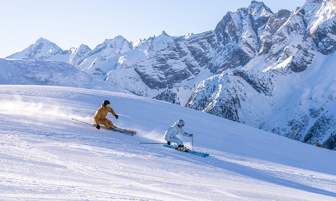 3. Adventtürchen - Abeneuer im Schnee Zwei Skifahrer gleiten auf einer Piste vor einem beeindruckenden, schneebedeckten Bergpanorama. Links fährt eine Person in brauner/gelber Kleidung, rechts eine Person in hellgrau/weißer Kleidung. Viel Schnee staubt auf, der Himmel ist hellblau.