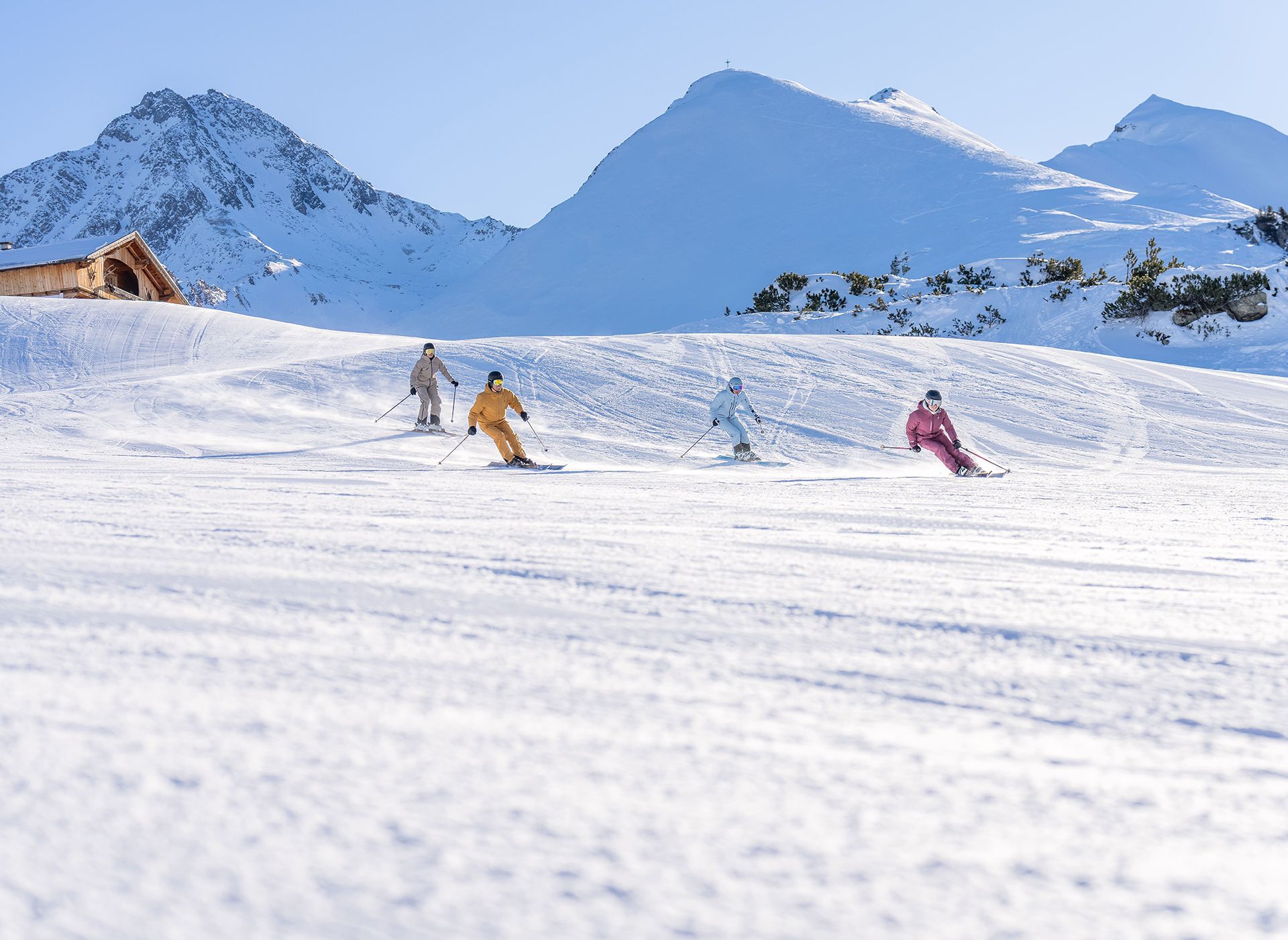 Vier Skifahrende in bunter Kleidung fahren auf einer weiten, verschneiten Piste. Im Hintergrund sind schneebedeckte Berge und ein kleines Holzhaus zu sehen.