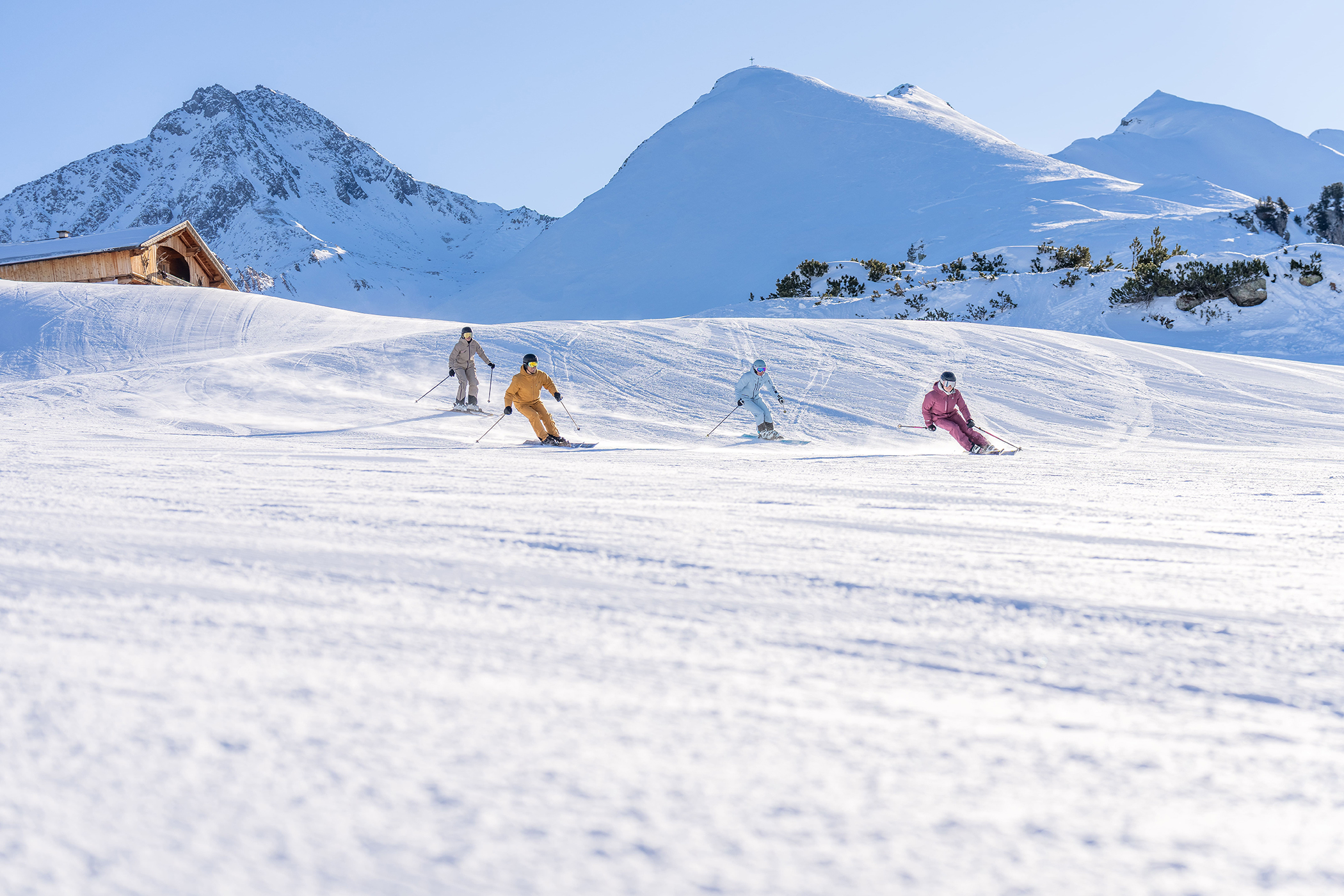 Four skiers in colorful clothing are skiing down a wide, snow-covered slope. Snow-capped mountains and a small wooden house can be seen in the background.