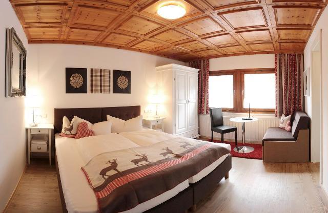 The picture shows a room with a wooden beam ceiling, a bed, and a seating area consisting of a bench, table, and chair. Decorative pictures hang on the wall, and the room has a window that lets in natural light.