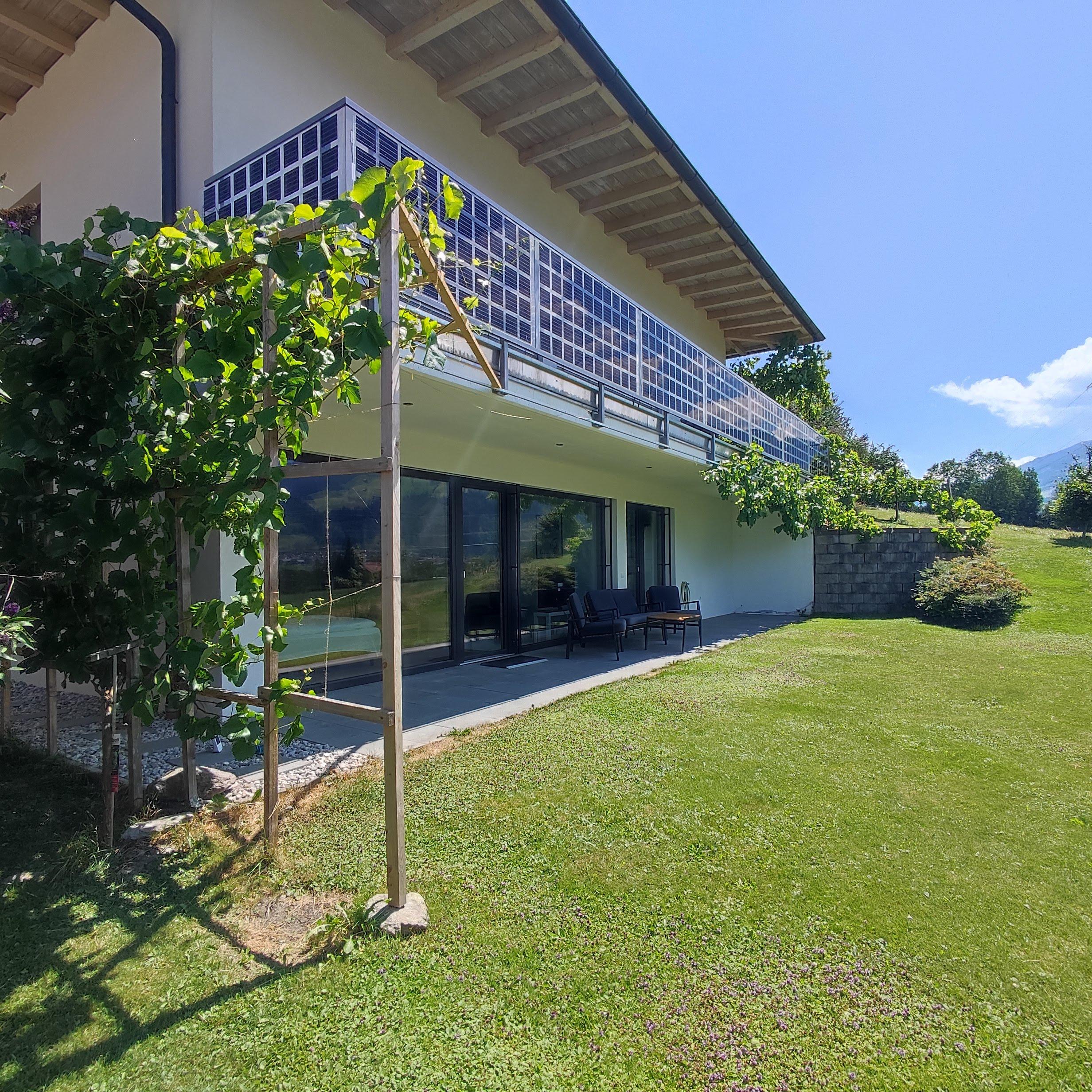 Modern balcony with glass railing, surrounded by greenery and garden, under a blue sky.