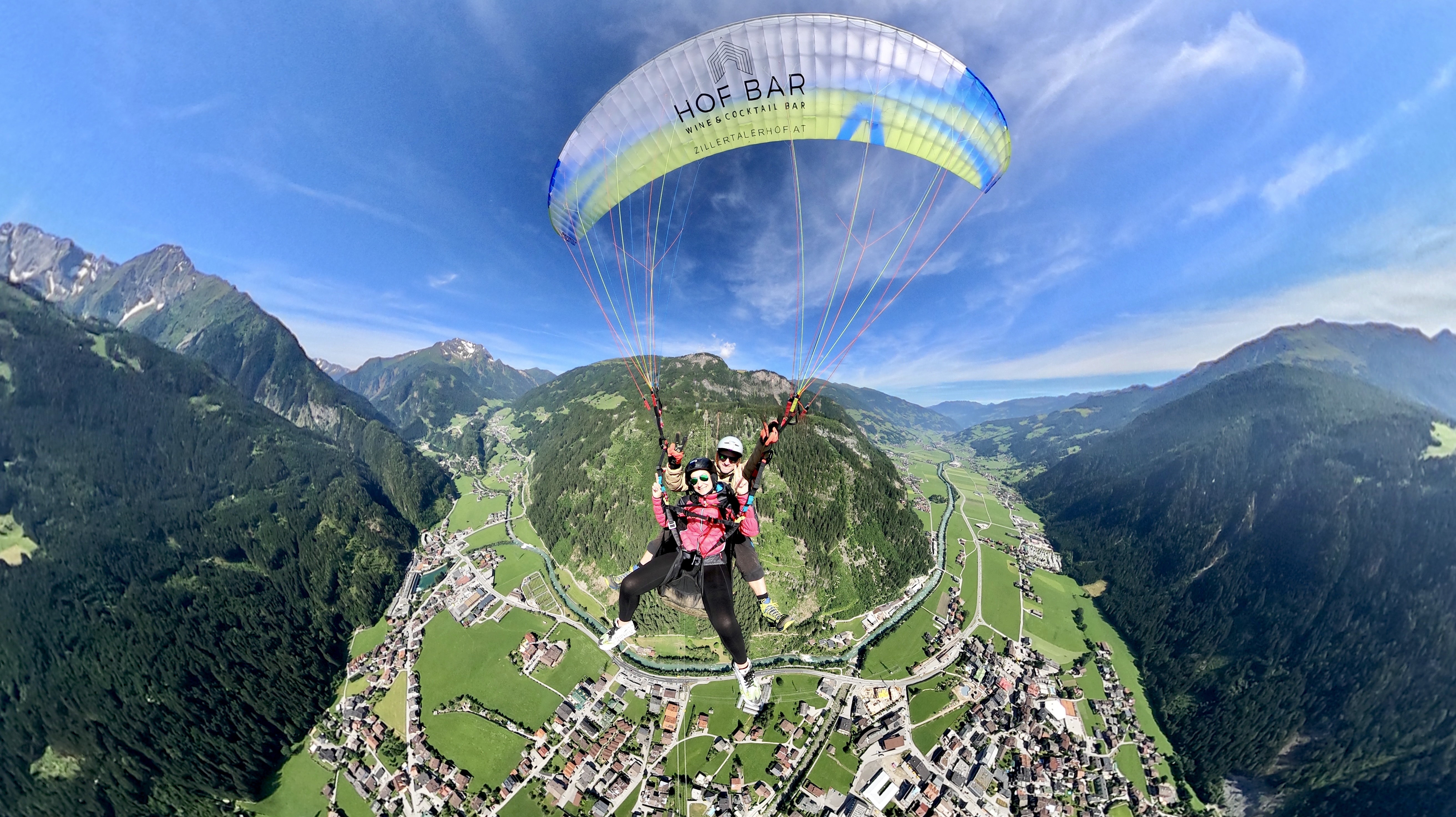 Das Bild zeigt zwei Personen, die mit einem Paragleiter über das Zillertal fliegen. Im Hintergrund sind beeindruckende Berge zu sehen, darüber erstreckt sich ein strahlend blauer Himmel mit ein paar weißen Wolken. 