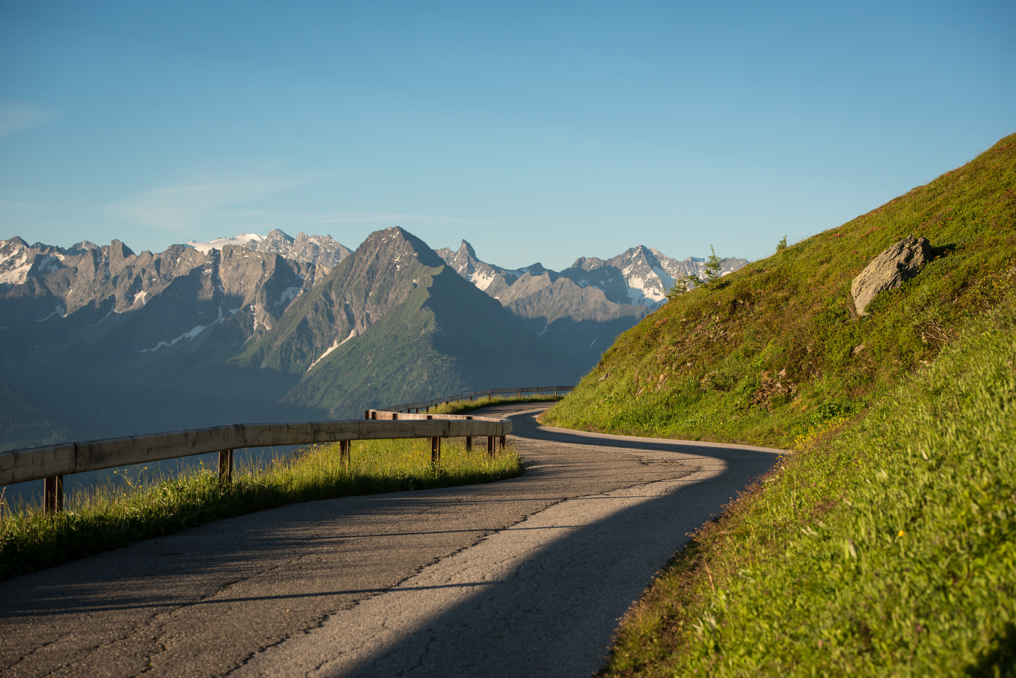 Sommerliche Aussicht auf die Zillertaler Höhenstraße: Eine kurvige Bergstraße führt durch grüne Almwiesen. Im Hintergrund erstreckt sich ein beeindruckendes Alpenpanorama mit schneebedeckten Gipfeln unter klarem Himmel.