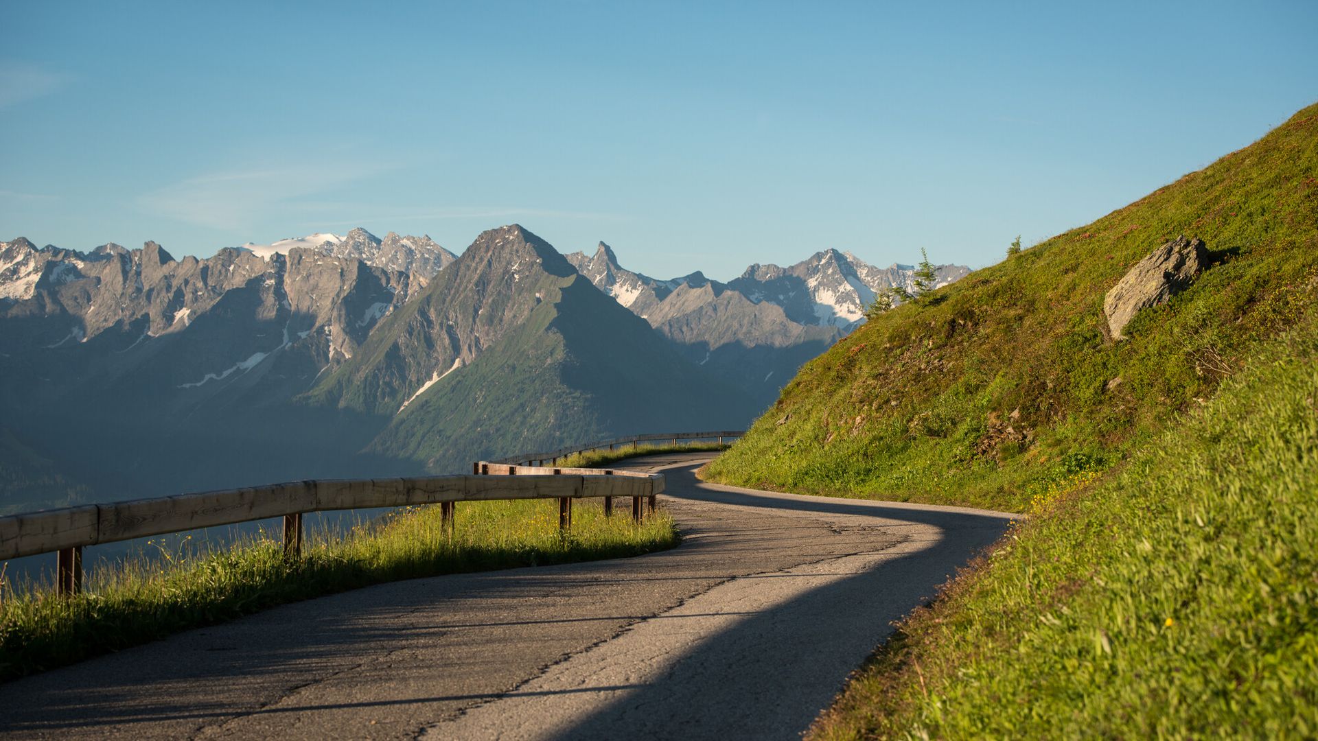 Summer view of the Zillertal High Alpine Road: A winding mountain road runs through green alpine meadows. In the background, a stunning panorama of snow-covered peaks stretches beneath a clear sky.
