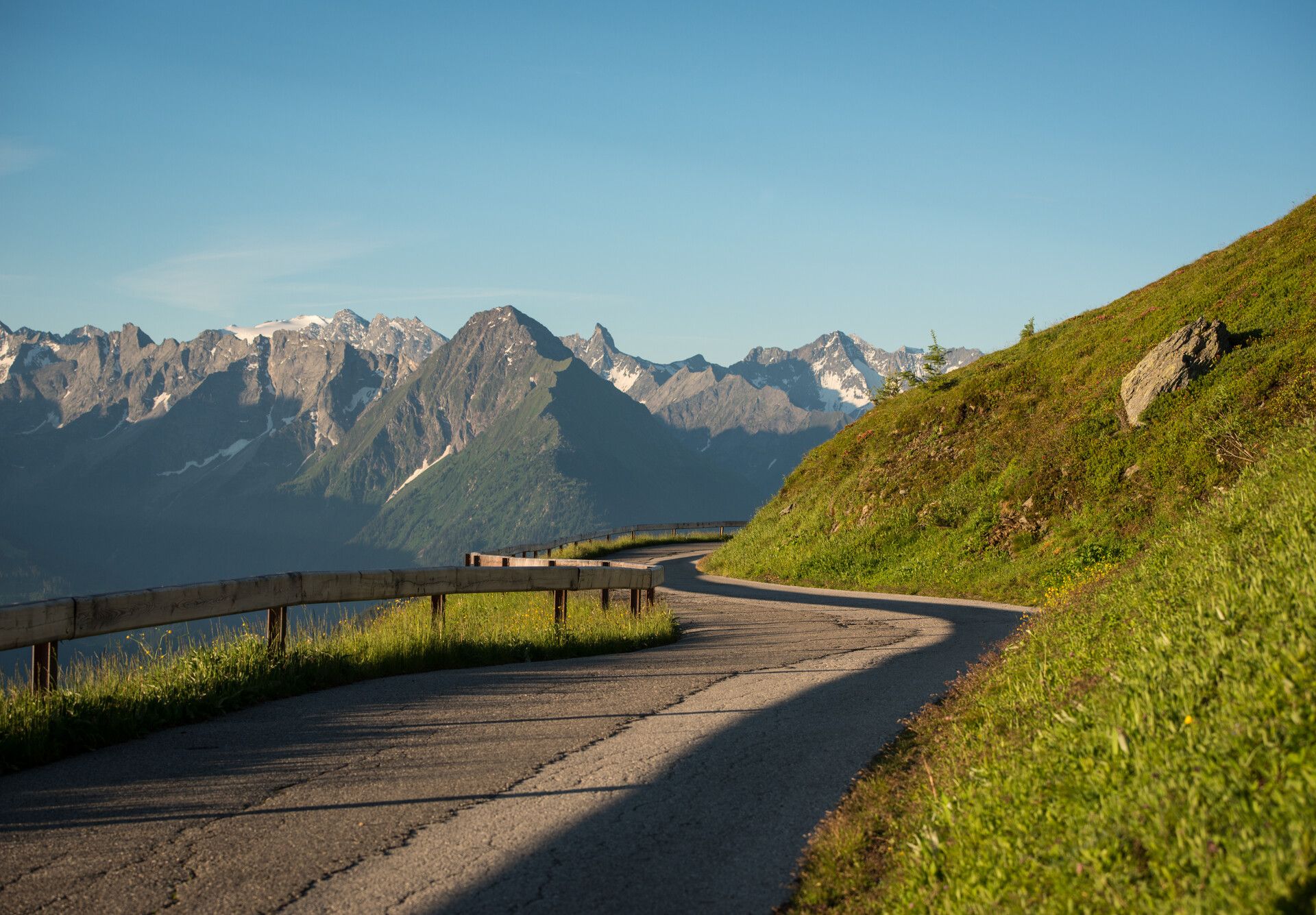 Summer view of the Zillertal High Alpine Road: A winding mountain road runs through green alpine meadows. In the background, a stunning panorama of snow-covered peaks stretches beneath a clear sky.
