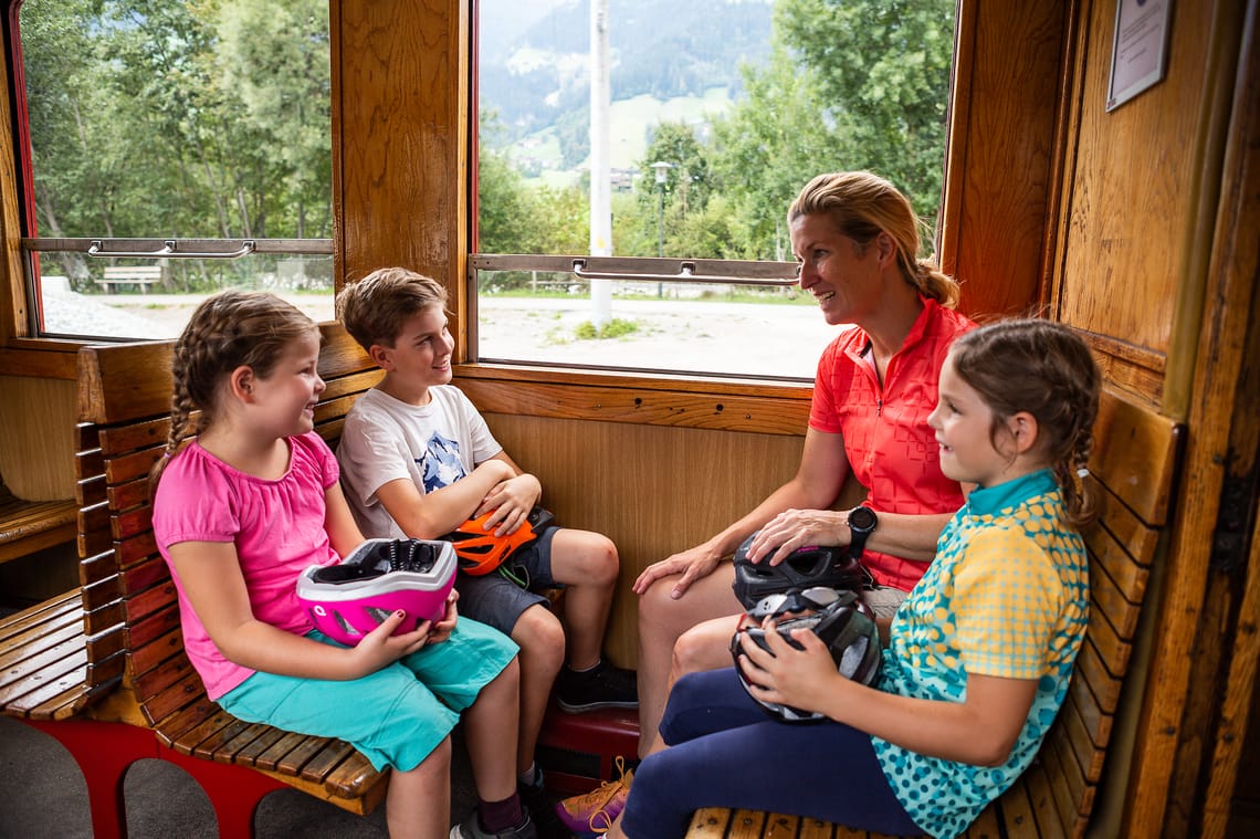 A ride on the Zillertalbahn is an experience for young and old. Three children and an adult are sitting on wooden benches on the Zillertalbahn. They are holding bicycle helmets in their hands and chatting. The green landscape of the Zillertal Valley can be seen through the window.