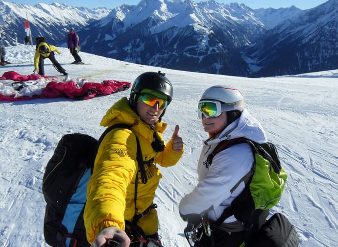 Zwei Skifahrer in Winterkleidung auf verschneitem Berg mit Bergen im Hintergrund, einer gibt Daumen hoch.