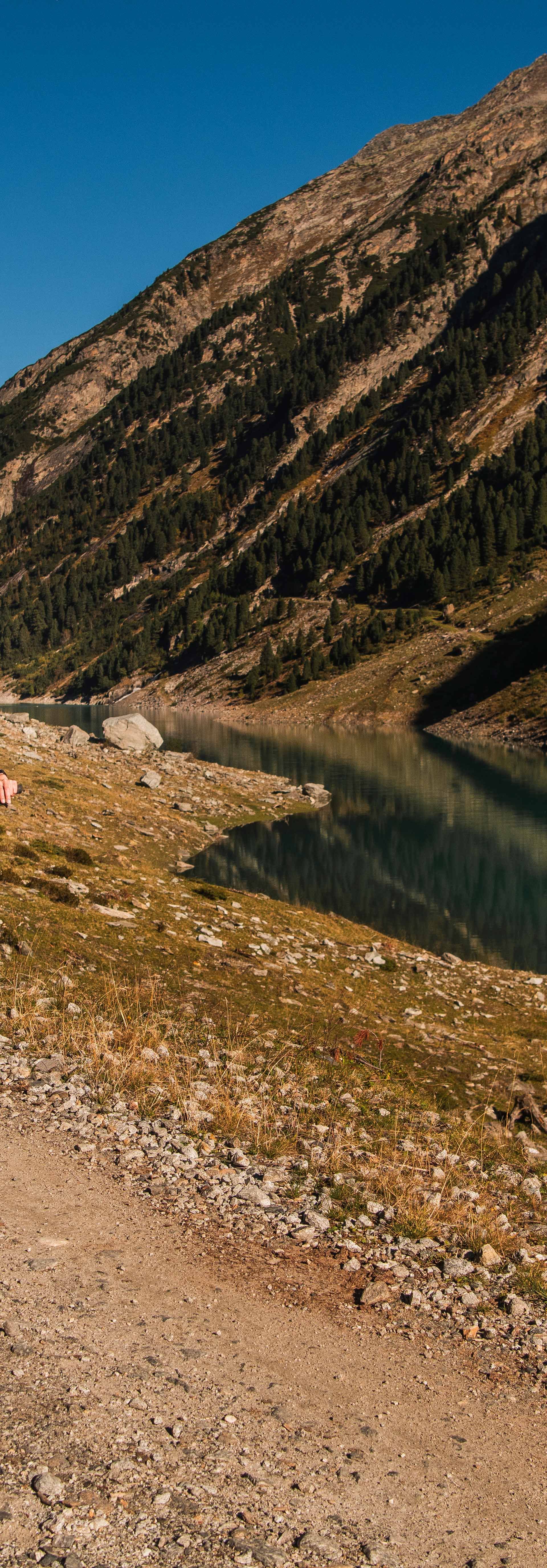 Bike and hike in the Zillertal: two people ride mountain bikes along a gravel path beside the Schlegeisspeicher near Mayrhofen-Hippach; turquoise water, steep slopes and alpine scenery in the background.