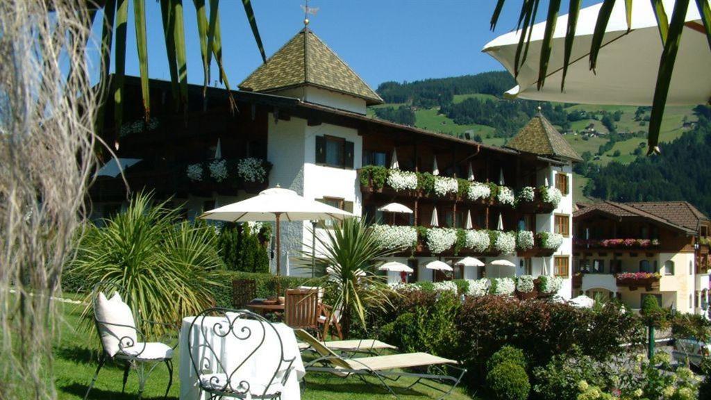 Das Bild zeigt das Hotel Ferienschlössl Alpenblick von außen. Im Vordergrund ist ein Garten mit einem Sonnenschirm zu sehen. Im Hintergrund ragen beeindruckende Berge unter einem klaren blauen Himmel empor.