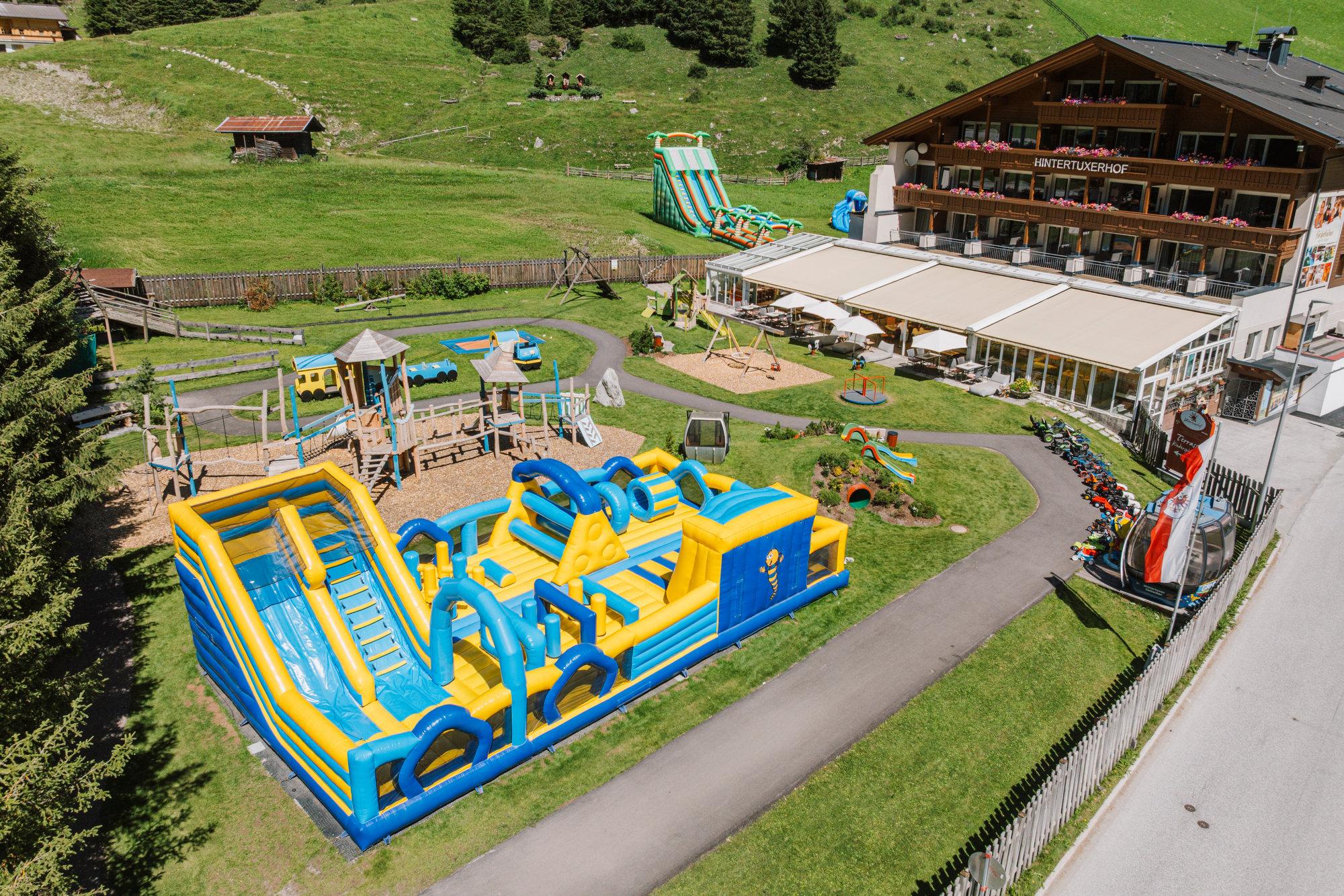 Blick auf einen Spielplatz mit Wasser- und Kletteranlagen, umgeben von einem Hotel im Alpenstil, auf grüner Wiese.