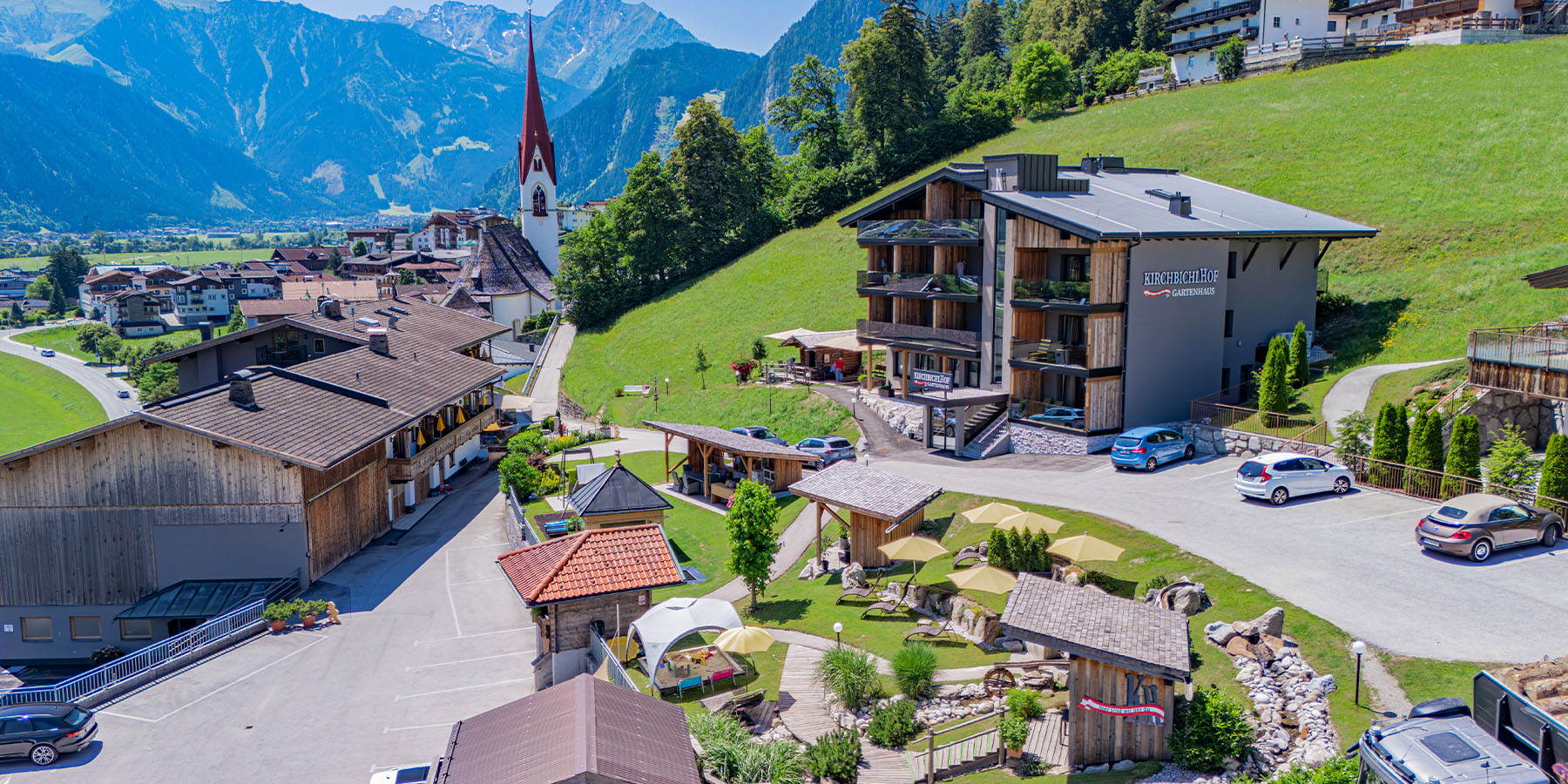 Aerial view of Hippach in the Zillertal: modern hotel with wooden facade and balcony, traditional village buildings, church steeple with red roof and snow-capped mountain peaks in the background in bright sunshine.