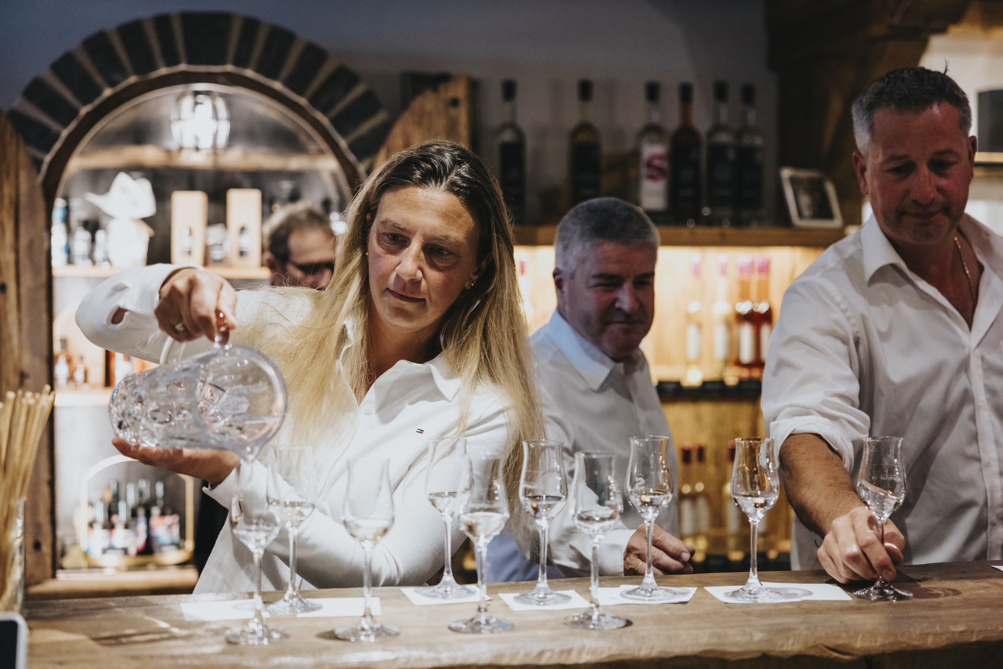 Eine Brennmeisterin schenkt „Maysterwurz“ in Gläser ein, während zwei Männer im Hintergrund an einer Bar stehen. Im Regal hinter ihnen reihen sich Flaschen.