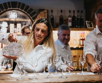 Eine Brennmeisterin schenkt „Maysterwurz“ in Gläser ein, während zwei Männer im Hintergrund an einer Bar stehen. Im Regal hinter ihnen reihen sich Flaschen.
