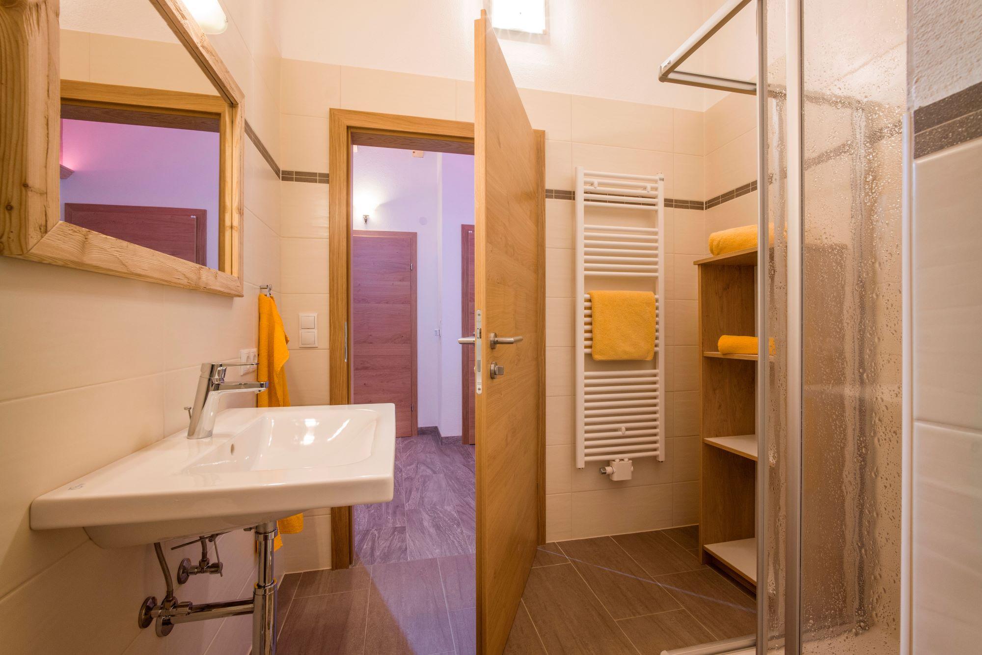 Badezimmer mit Waschbecken, Handtüchern, Spiegel, Heizkörper und Dusche, alles in warmen Holztönen und beige Fliesen.