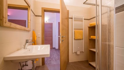 Badezimmer mit Waschbecken, Handtüchern, Spiegel, Heizkörper und Dusche, alles in warmen Holztönen und beige Fliesen.