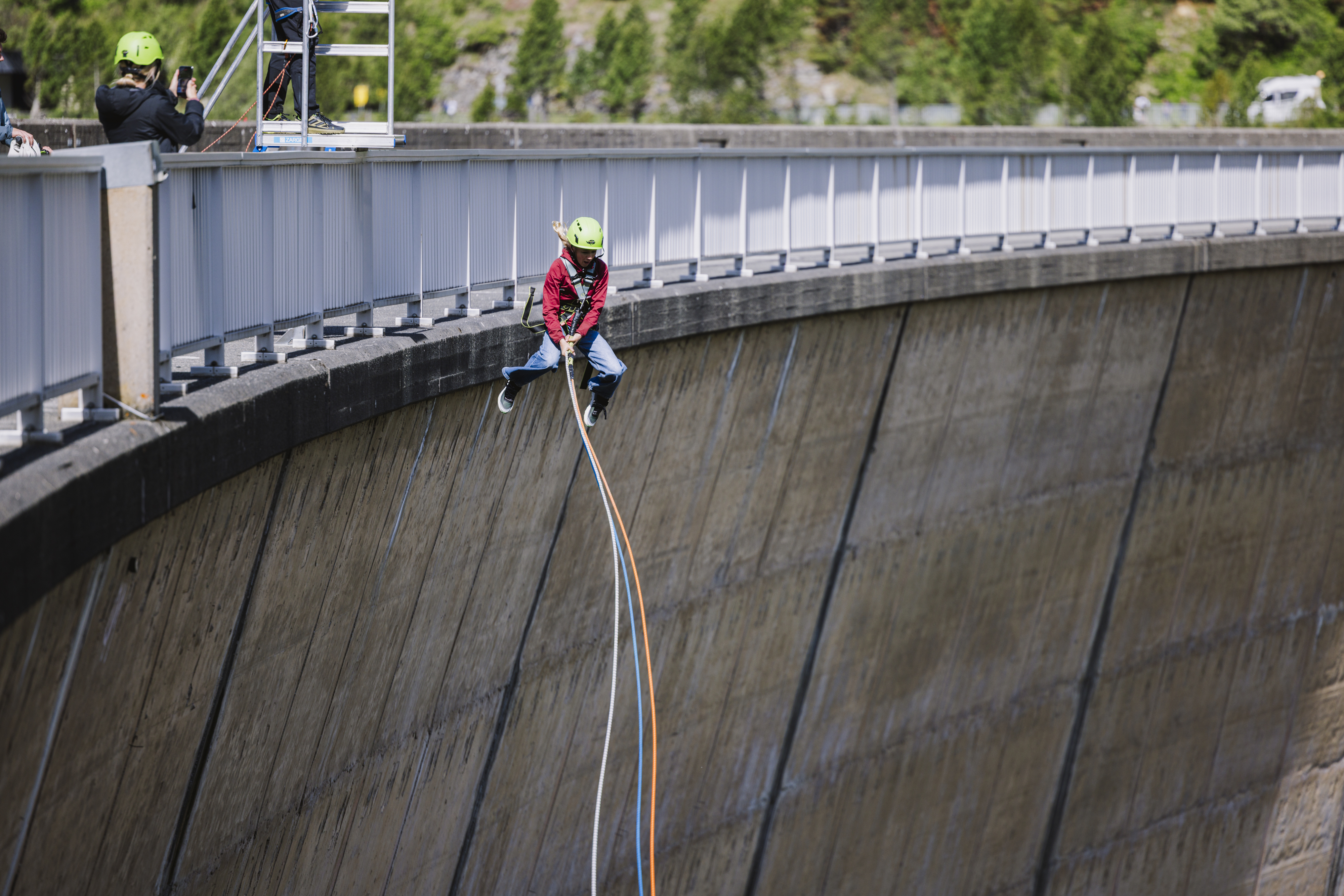 Child with helmet and harness swings on a rope over a dam, others watch and take photos.