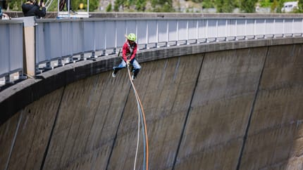 Child with helmet and harness swings on a rope over a dam, others watch and take photos.