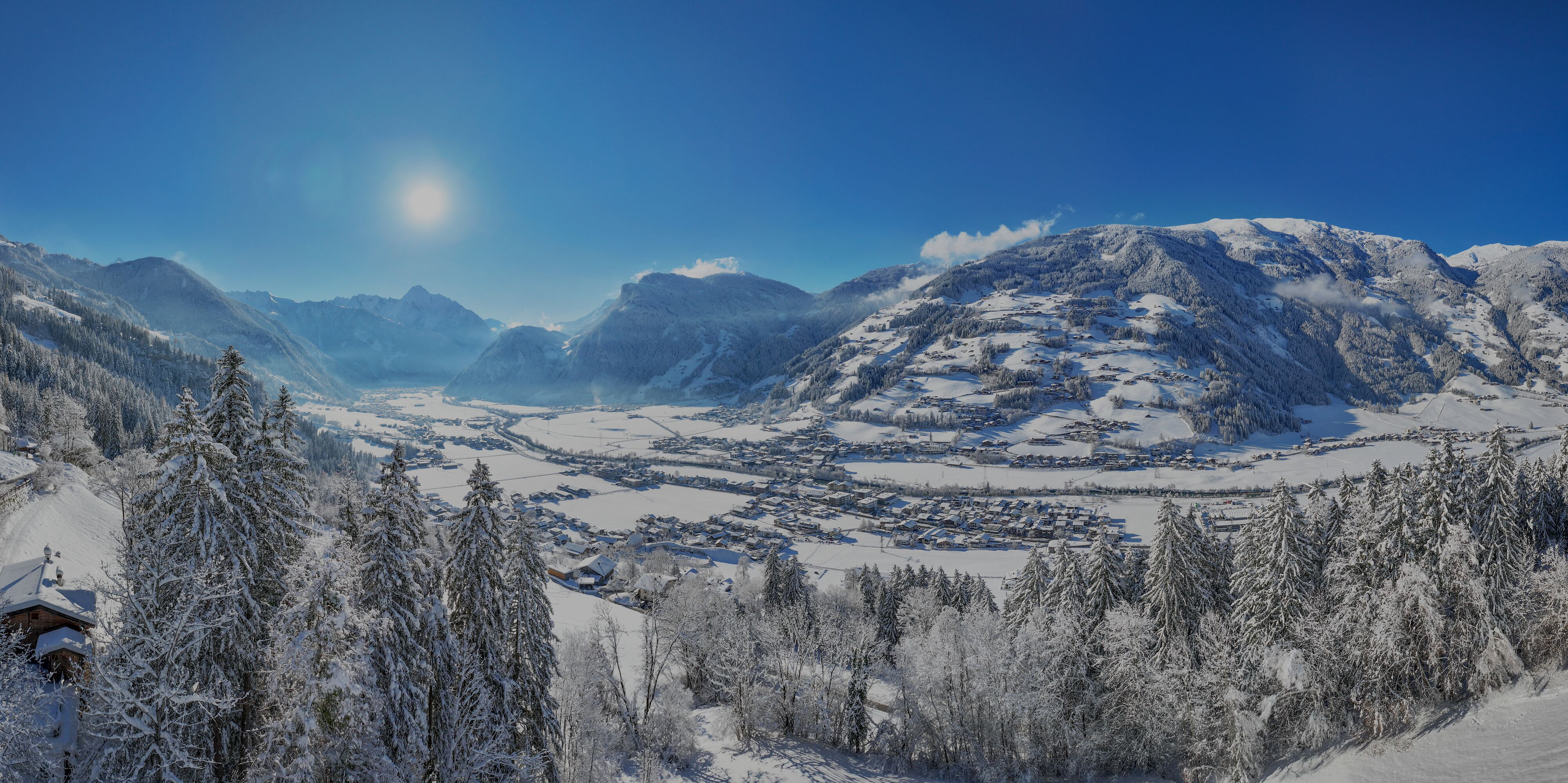 Panoramablick ins hintere Zillertal im Winter: verschneites Tal mit Mayrhofen/Hippach, Tannenwald im Vordergrund, Sonnenschein und schneebedeckte Alpen unter blauem Himmel.