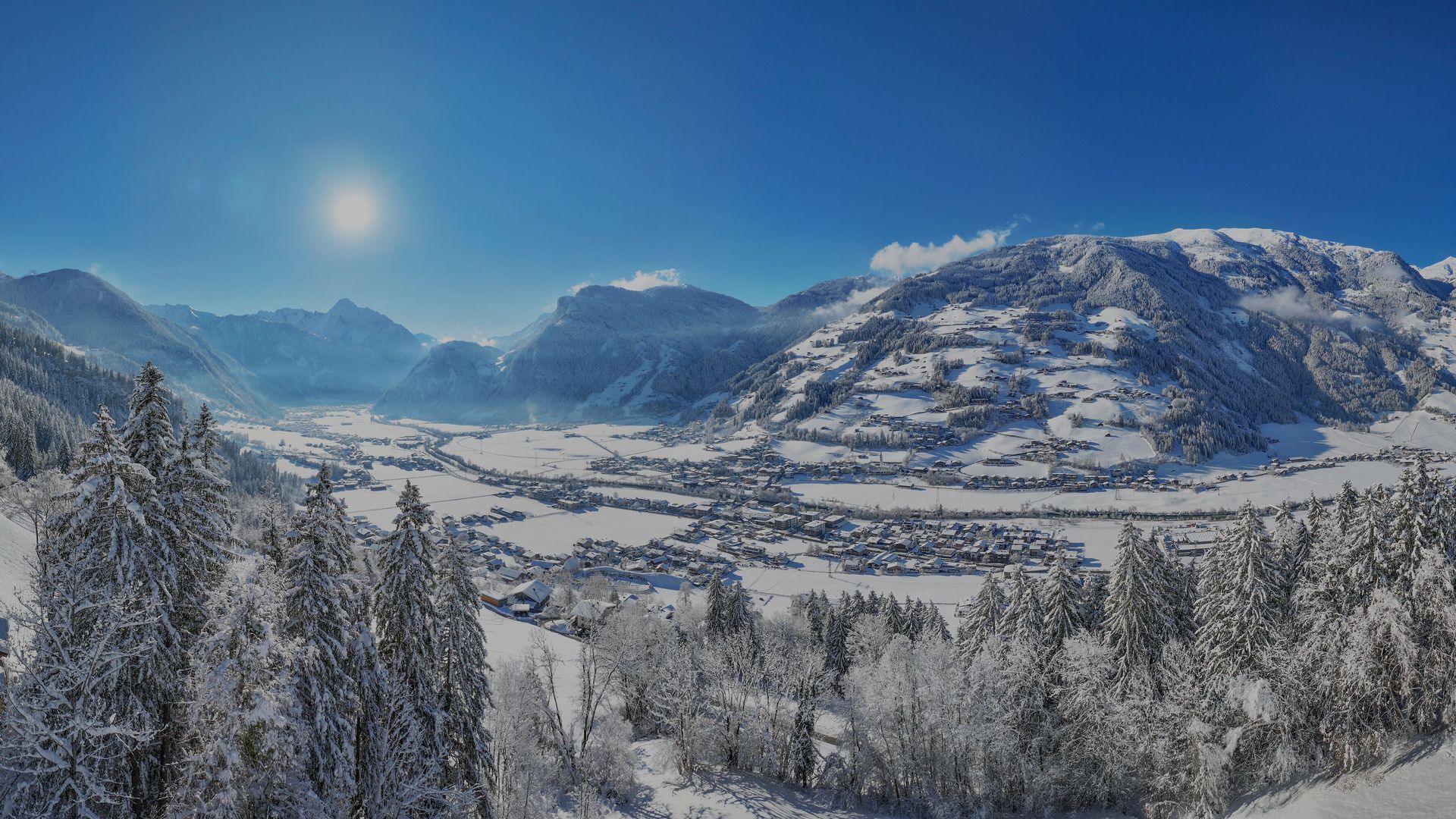 Panoramablick ins hintere Zillertal im Winter: verschneites Tal mit Mayrhofen/Hippach, Tannenwald im Vordergrund, Sonnenschein und schneebedeckte Alpen unter blauem Himmel.
