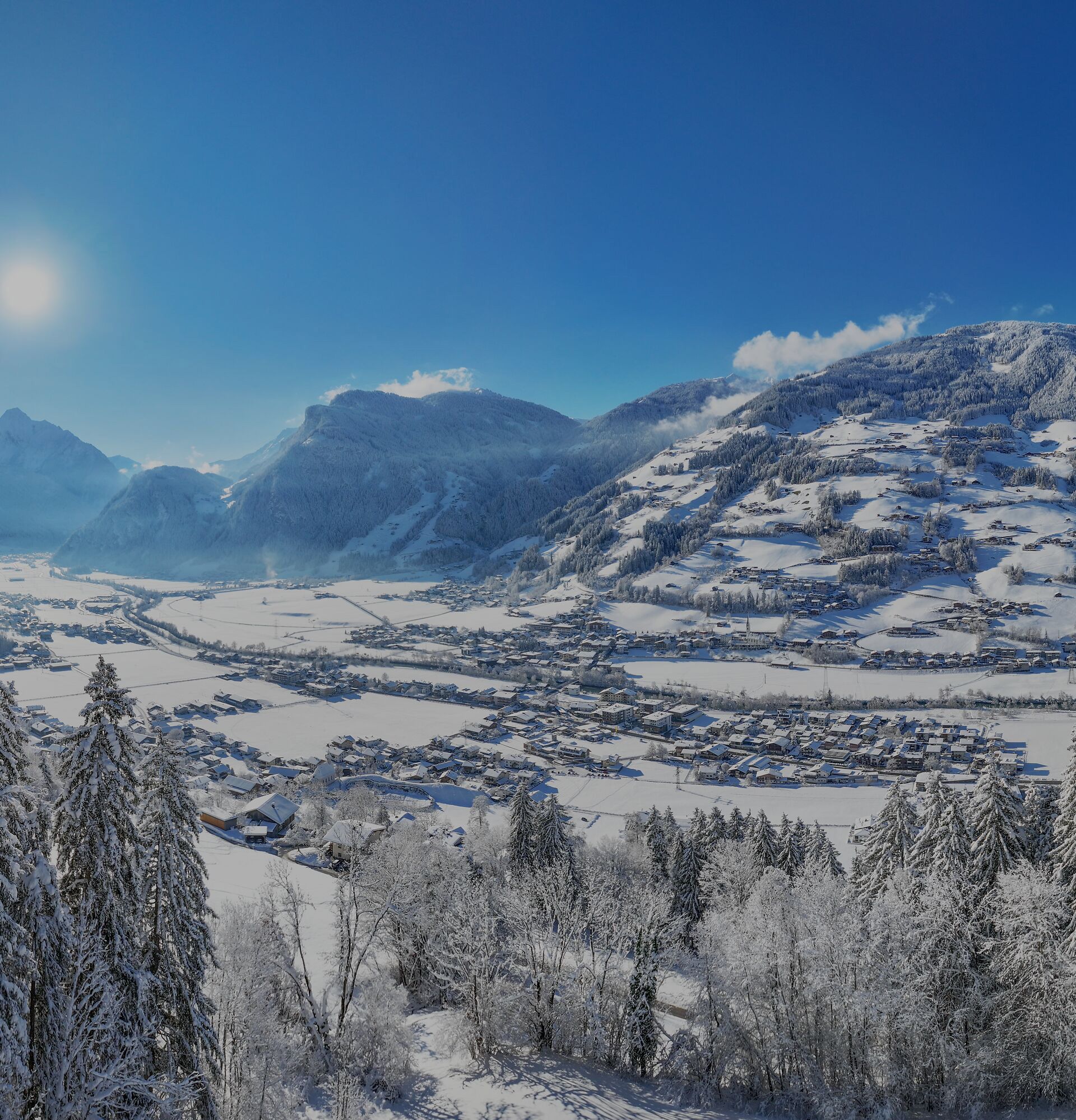 Panoramablick ins hintere Zillertal im Winter: verschneites Tal mit Mayrhofen/Hippach, Tannenwald im Vordergrund, Sonnenschein und schneebedeckte Alpen unter blauem Himmel.