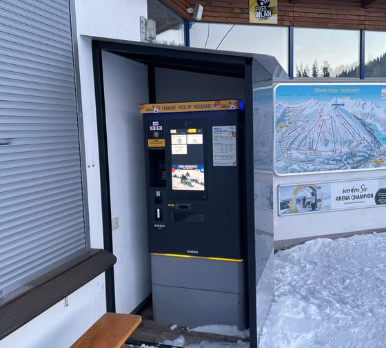 Ski lift ticket machine outside a mountain hut, with snow on the ground and a bench nearby.