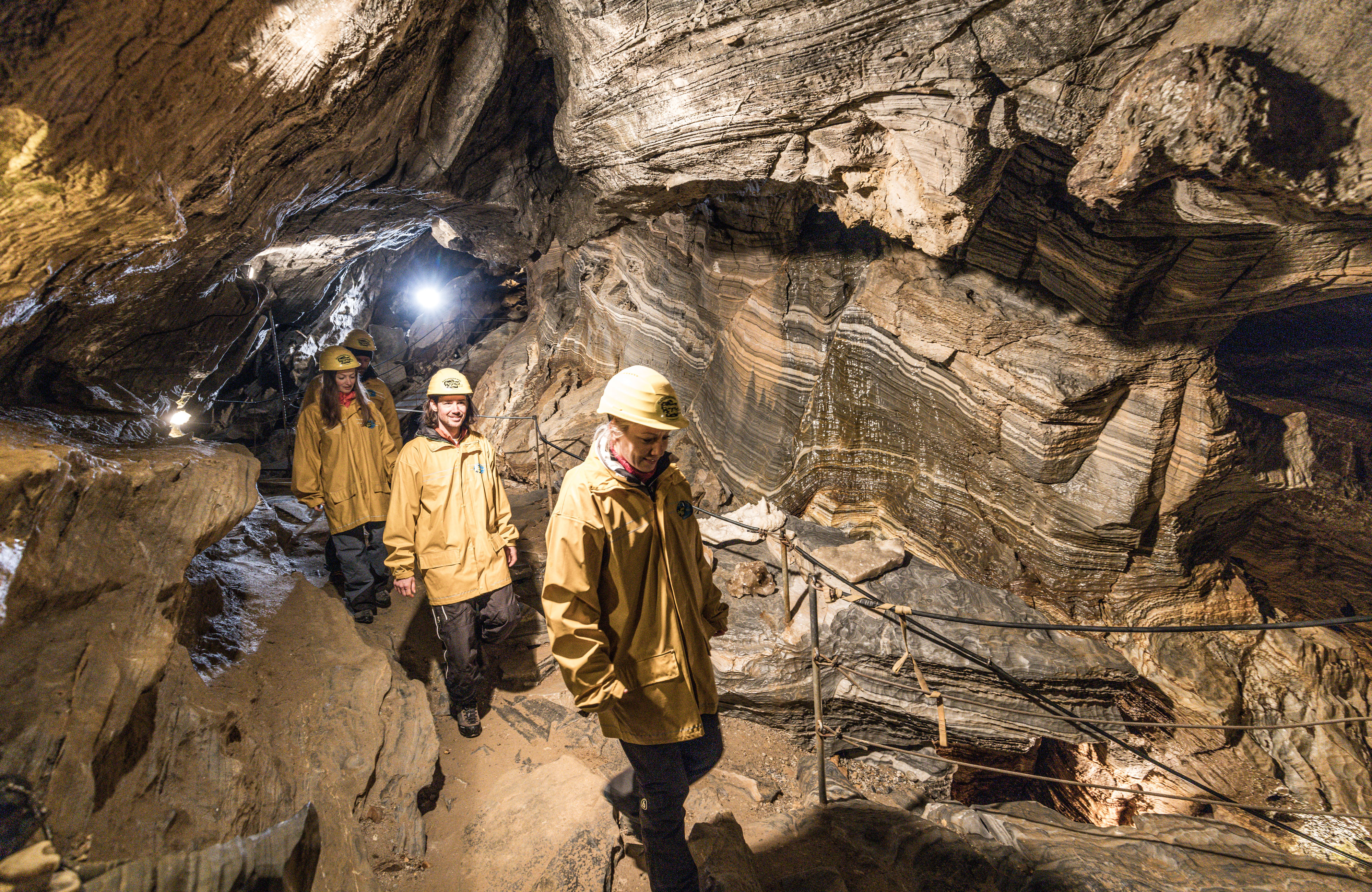 Besucher mit Helmen und Helmlampen gehen durch die Spannagelhöhle am Hintertuxer Gletscher, das Licht der Lampen beleuchtet die rauen Felswände des Naturdenkmals, der Weg führt über unebenen Felsboden.