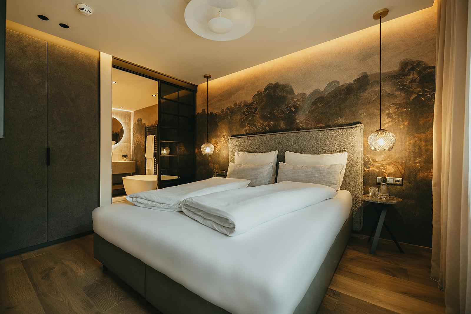 Modernes Schlafzimmer mit Wandbild, beigem Kopfteil, Nachttisch, Hängelampen und Blick ins Bad.