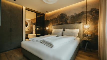 Modernes Schlafzimmer mit Wandbild, beigem Kopfteil, Nachttisch, Hängelampen und Blick ins Bad.