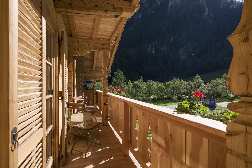 Holzveranda mit Stühlen, Blick auf grüne Bäume und Berge im Hintergrund, blühende Geranien auf dem Geländer.