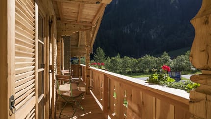 Holzveranda mit Stühlen, Blick auf grüne Bäume und Berge im Hintergrund, blühende Geranien auf dem Geländer.