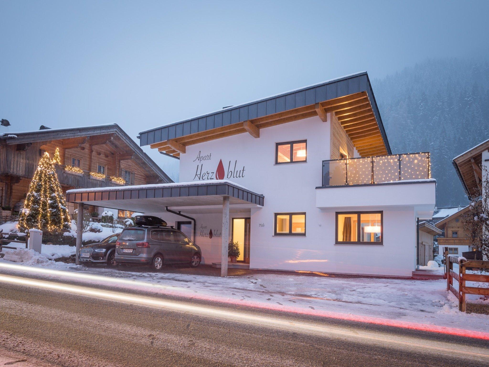 Modern house with wood and white exterior, Christmas lights, and snow-covered roof in mountain area.
