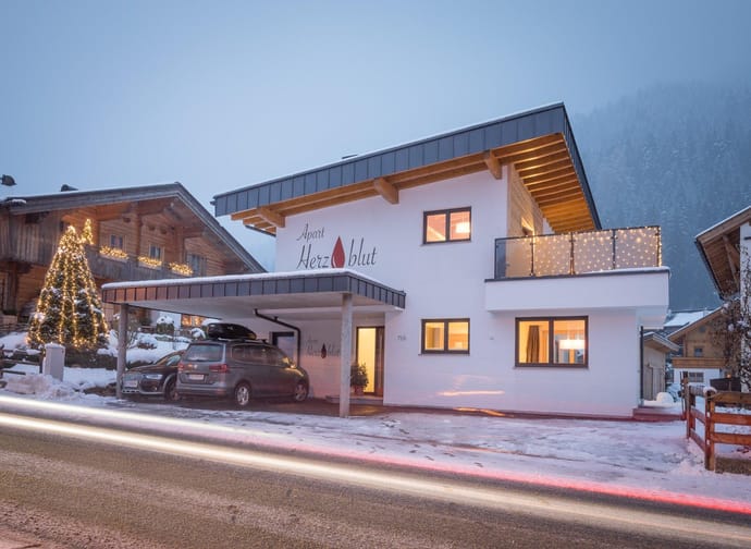 Modern house with wood and white exterior, Christmas lights, and snow-covered roof in mountain area.