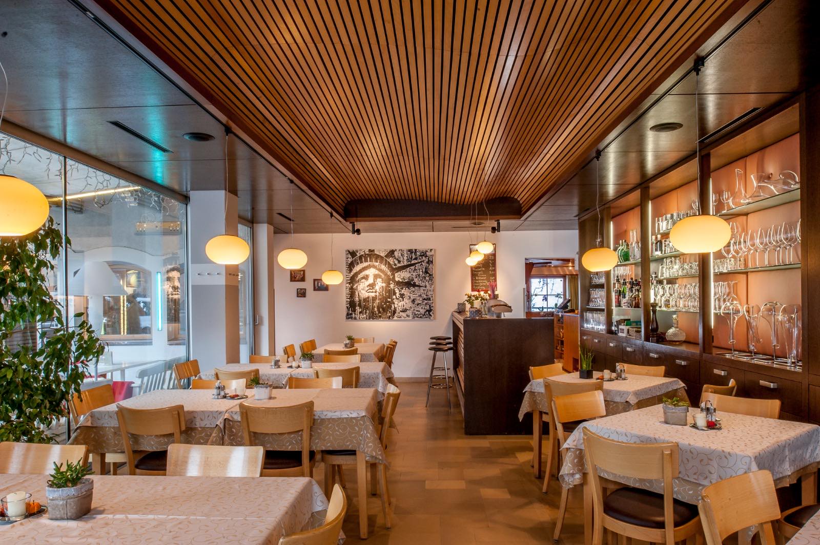 Das Bild zeigt das Café Tirol von innen. Der Raum wirkt sehr gemütlich mit Holzelementen, stilvollen Lampen und einladend gedeckten Tischen. Große Fenster lassen viel Tageslicht herein und bieten einen schönen Ausblick.