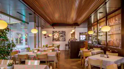 Das Bild zeigt das Café Tirol von innen. Der Raum wirkt sehr gemütlich mit Holzelementen, stilvollen Lampen und einladend gedeckten Tischen. Große Fenster lassen viel Tageslicht herein und bieten einen schönen Ausblick.