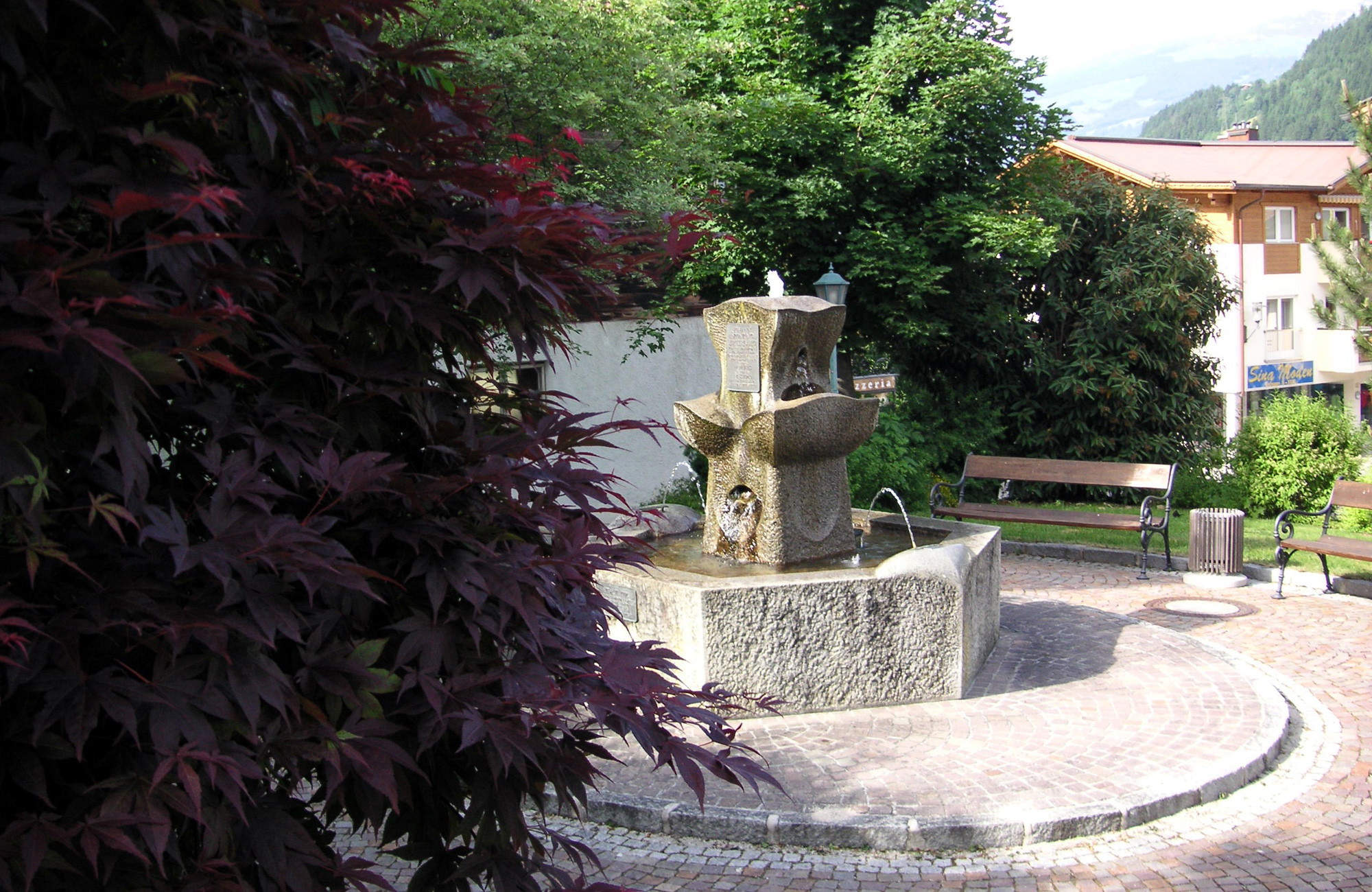 Johann Sponring stone fountain surrounded by trees, benches, and houses on the Schwendau Trail in Tyrol, Station 13 “Johann Sponring Fountain,” photographed by Paul Wechselberger.