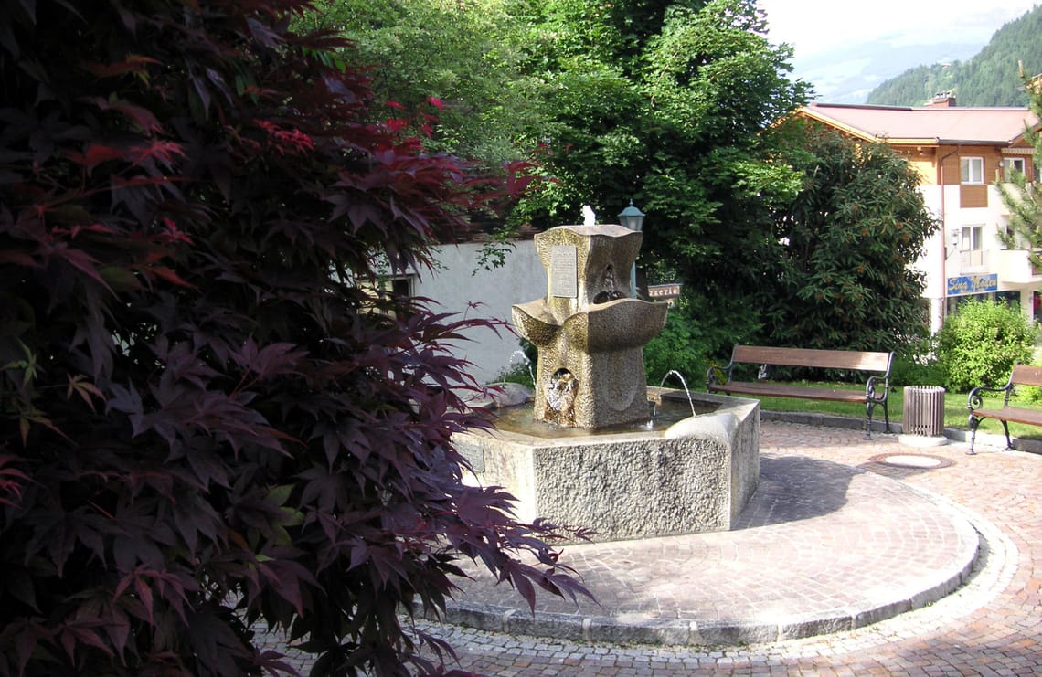 Schwendauer Pfad - Johann-Sponring-Brunnen Johann-Sponring-Brunnen aus Stein, umgeben von Bäumen, Bänken und Häusern, am Schwendauer Pfad in Tirol, Station 13 „Johann-Sponring-Brunnen“, fotografiert von Paul Wechselberger.
