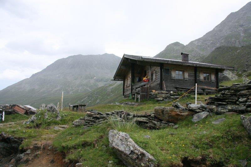 A wooden cabin on a grassy, rocky hillside with mountains in the background.