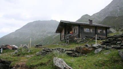A wooden cabin on a grassy, rocky hillside with mountains in the background.