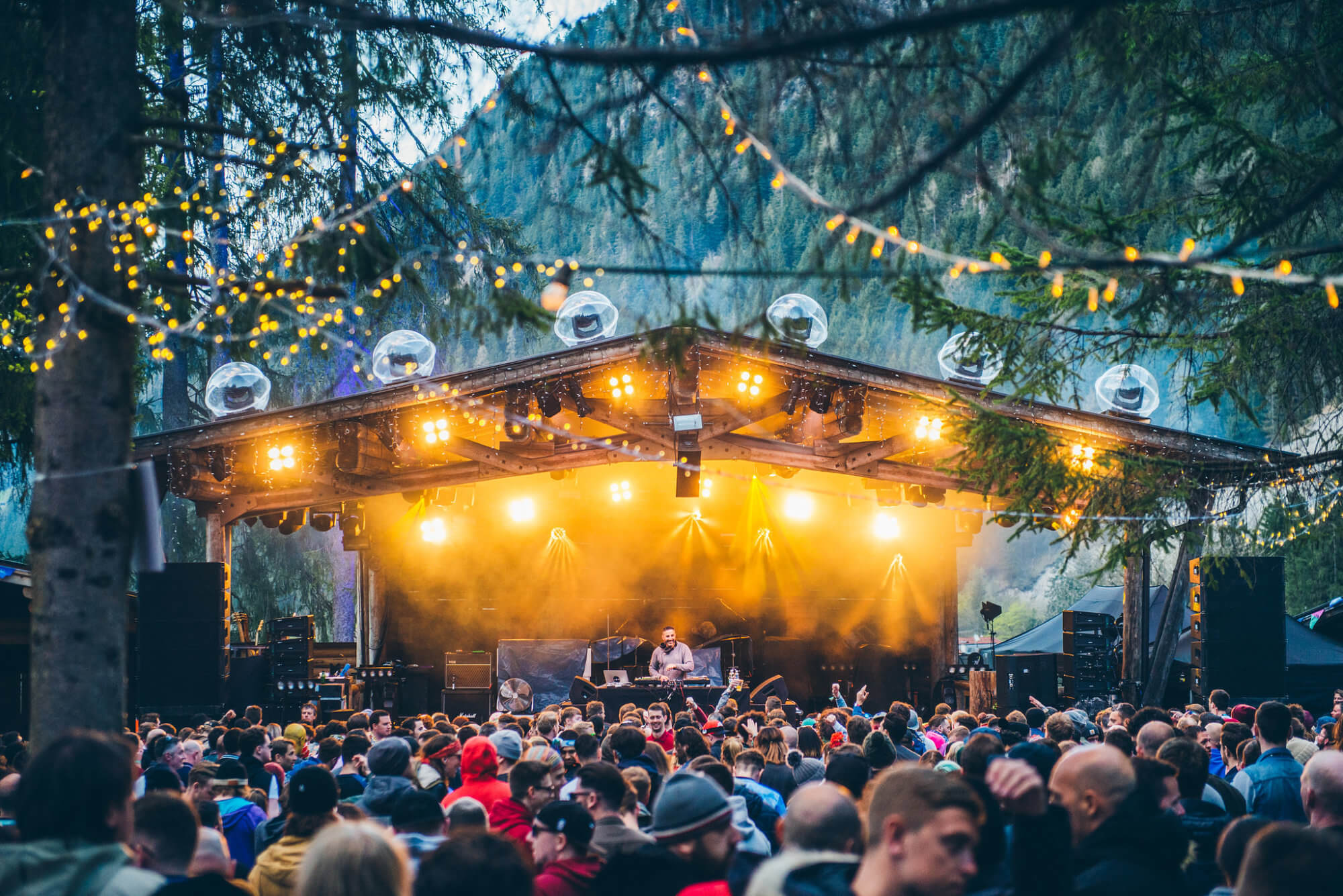 The image shows a vibrant open-air party in the forest with an illuminated stage, a DJ, and a crowd celebrating under sparkling string lights.