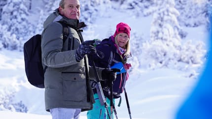 The image shows two people winter hiking in a heavily snow-covered terrain. They wear warm winter clothing, backpacks, and use trekking poles. In the background, snow-covered trees and a clear sky can be seen, creating an idyllic winter landscape.
